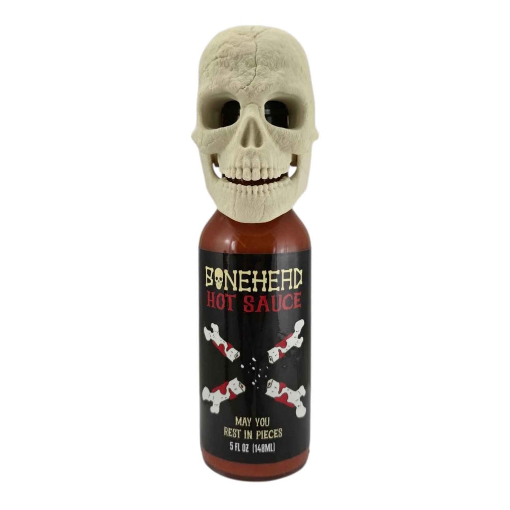 A 5-fl-oz bottle of Bone Head Hot Sauce with a black label featuring bone graphics and red lettering. The bottle is topped with a detailed 3D-printed beige skull with an articulated jaw. The label reads āBone Head Hot Sauce ā May You Rest in Pieces.ā UPC 701705500344.