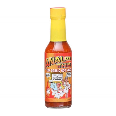 A bottle of ANALize This XXX Garlic Hot Sauce, featuring a bold and eye-catching label. The hot sauce is marketed as a spicy condiment that emphasizes both flavor and digestive health. It contains habanero peppers, fresh garlic, crushed tomatoes, key lime juice, and various other ingredients. The caption mentions its availability exclusively at PepperHotSpot.com and warns about the sauce's intense heat.