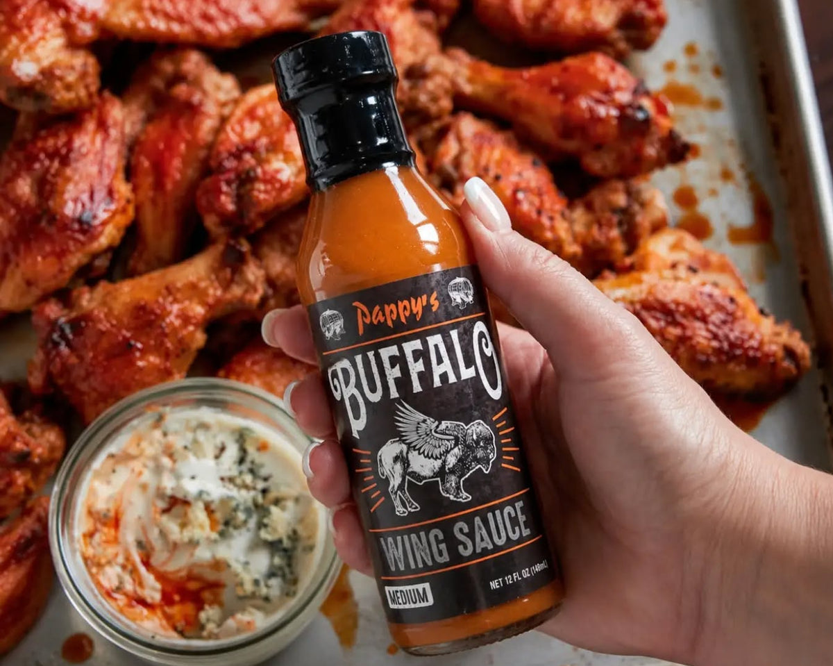 A hand holding a bottle of Pepper's Buffalo Wing Sauce in front of a tray of buffalo chicken wings and a small bowl of blue cheese dressing.