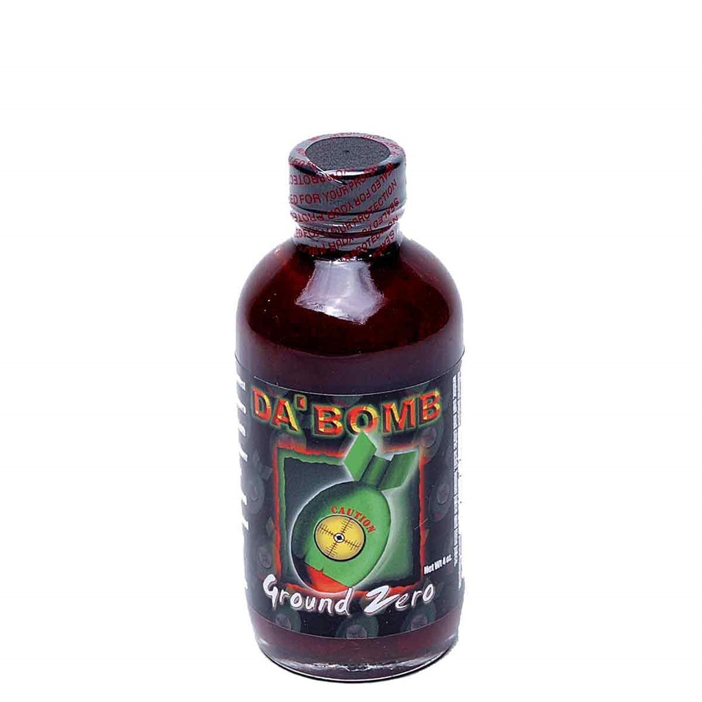 A dark glass bottle of Da’ Bomb “Ground Zero” Hot Sauce. The label shows a green bomb graphic with a yellow caution symbol in the center, set against a black background with subtle fiery tones. The text reads “Da’ Bomb” at the top and “Ground Zero” along the bottom. The cap is sealed with tamper-evident red tape printed with warning text.