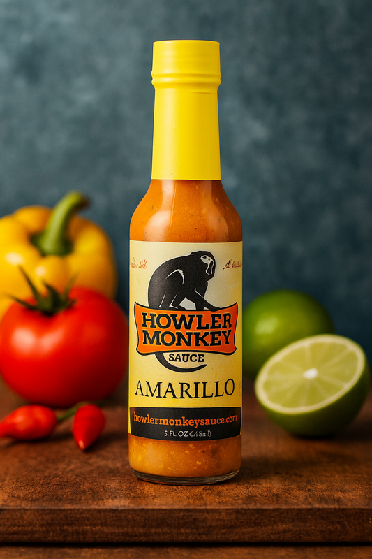 A bottle of Howler Monkey Amarillo Hot Sauce with a bright yellow cap and creamy orange sauce inside. The label features the black howler monkey logo and bold orange lettering. The bottle is set on a wooden surface with fresh ingredients in the background, including a tomato, yellow bell pepper, red chili peppers, and a sliced lime.