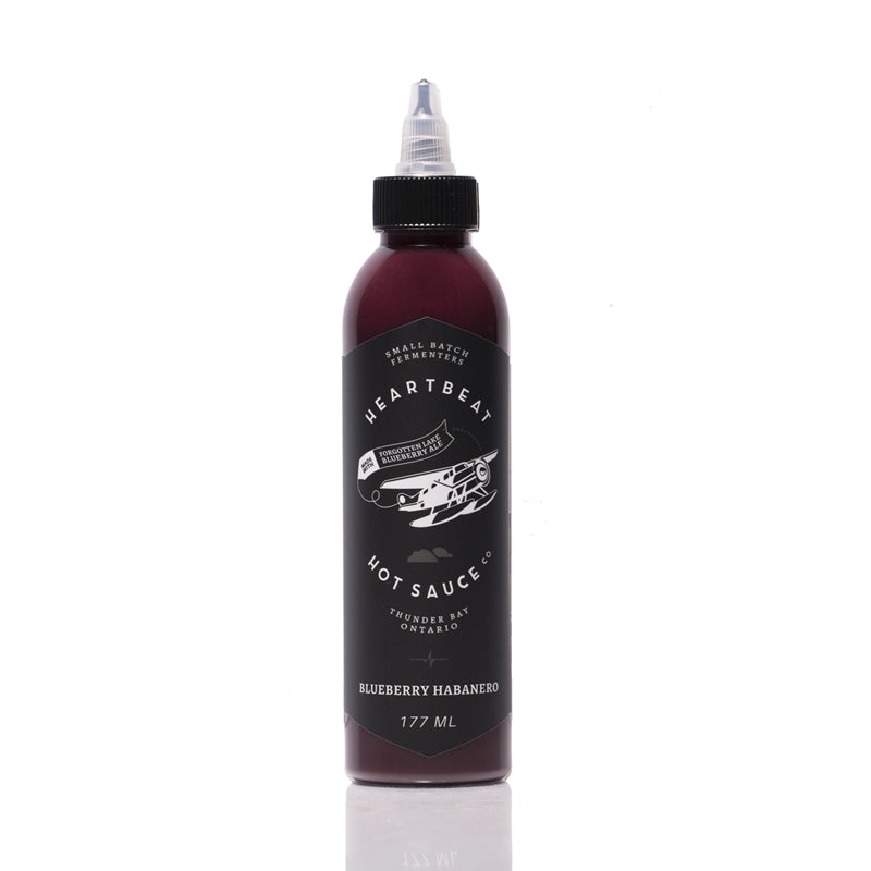 Tall squeeze bottle of Heartbeat Blueberry Habanero Hot Sauce with a black label featuring a white airplane graphic; deep purple sauce visible inside; labeled “Blueberry Habanero” with 177 mL noted at the bottom.