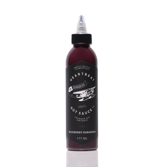 Tall squeeze bottle of Heartbeat Blueberry Habanero Hot Sauce with a black label featuring a white airplane graphic; deep purple sauce visible inside; labeled “Blueberry Habanero” with 177 mL noted at the bottom.