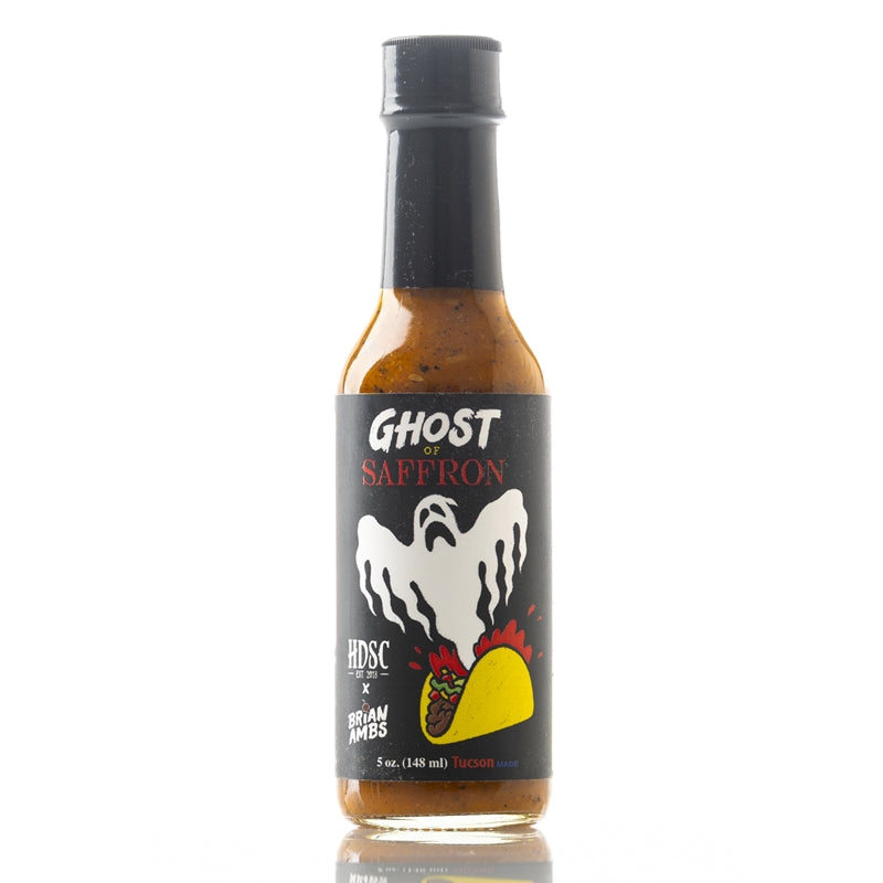 A clear glass bottle of High Desert Sauce Co. “Ghost of Saffron” Hot Sauce. The label is black with a white ghost rising from a yellow taco illustrated at the bottom, with red heat lines around it. The words “Ghost of Saffron” are printed in white and red text above the ghost. The bottle is filled with an orange-brown sauce containing visible spices, and it has a black cap.