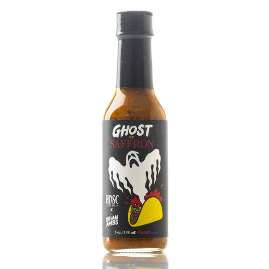 A clear glass bottle of High Desert Sauce Co. “Ghost of Saffron” Hot Sauce. The label is black with a white ghost rising from a yellow taco illustrated at the bottom, with red heat lines around it. The words “Ghost of Saffron” are printed in white and red text above the ghost. The bottle is filled with an orange-brown sauce containing visible spices, and it has a black cap.