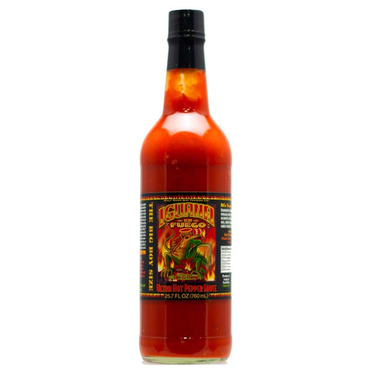 A tall 25.7 oz bottle of Iguana En Fuego Ultra Hot Pepper Sauce filled with bright orange-red sauce. The label features a colorful green iguana surrounded by flames and tropical artwork, with text indicating “Ultra Hot Pepper Sauce” and the large bottle size. UPC: 751059900796