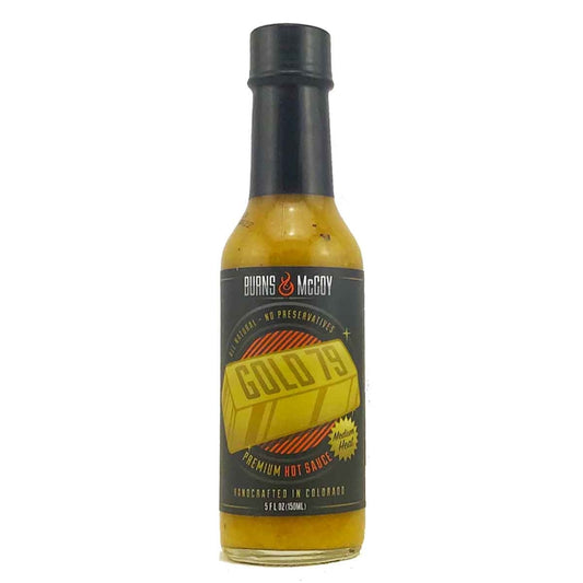 A bottle of Burns & McCoy Gold 79 Premium Hot Sauce. The bottle contains a yellow-gold sauce with visible seasoning flecks and has a black cap. The label is dark with a large illustrated gold bar in the center labeled “GOLD 79,” surrounded by text that reads “Burns & McCoy,” “All Natural – No Preservatives,” “Medium Heat,” and “Handcrafted in Colorado.”