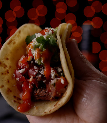 A close-up of a hand holding a soft corn taco filled with seasoned meat, diced red onion and tomato, guacamole, and hot sauce, set against a dark background with warm red bokeh lights.