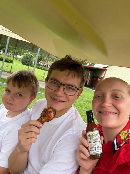Three people smiling under a beige canopy, with the center person holding a piece of grilled chicken and the person on the right holding a bottle of Dr Pepper Pepper Jack Hot Sauce.