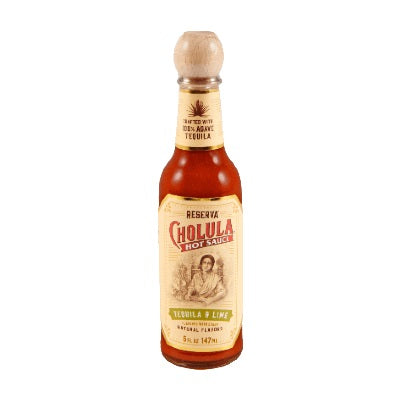 A bottle of Cholula Reserva Tequila & Lime Hot Sauce with an off-white label featuring a graphic of a woman and the Cholula logo. The sauce is visible inside the clear bottle and has a dark red color. The background is off-white with a banner at the top that reads "crafted with 100% agave tequila."